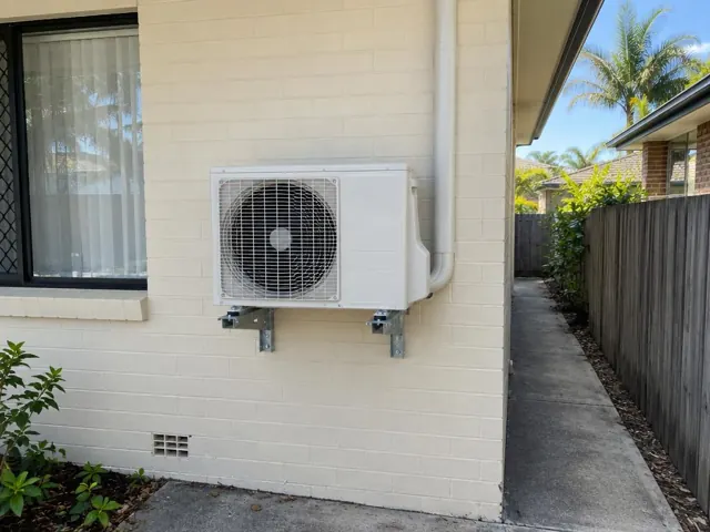 Wall-mounted outdoor air conditioner unit on a cream brick house beside a side path in Southport